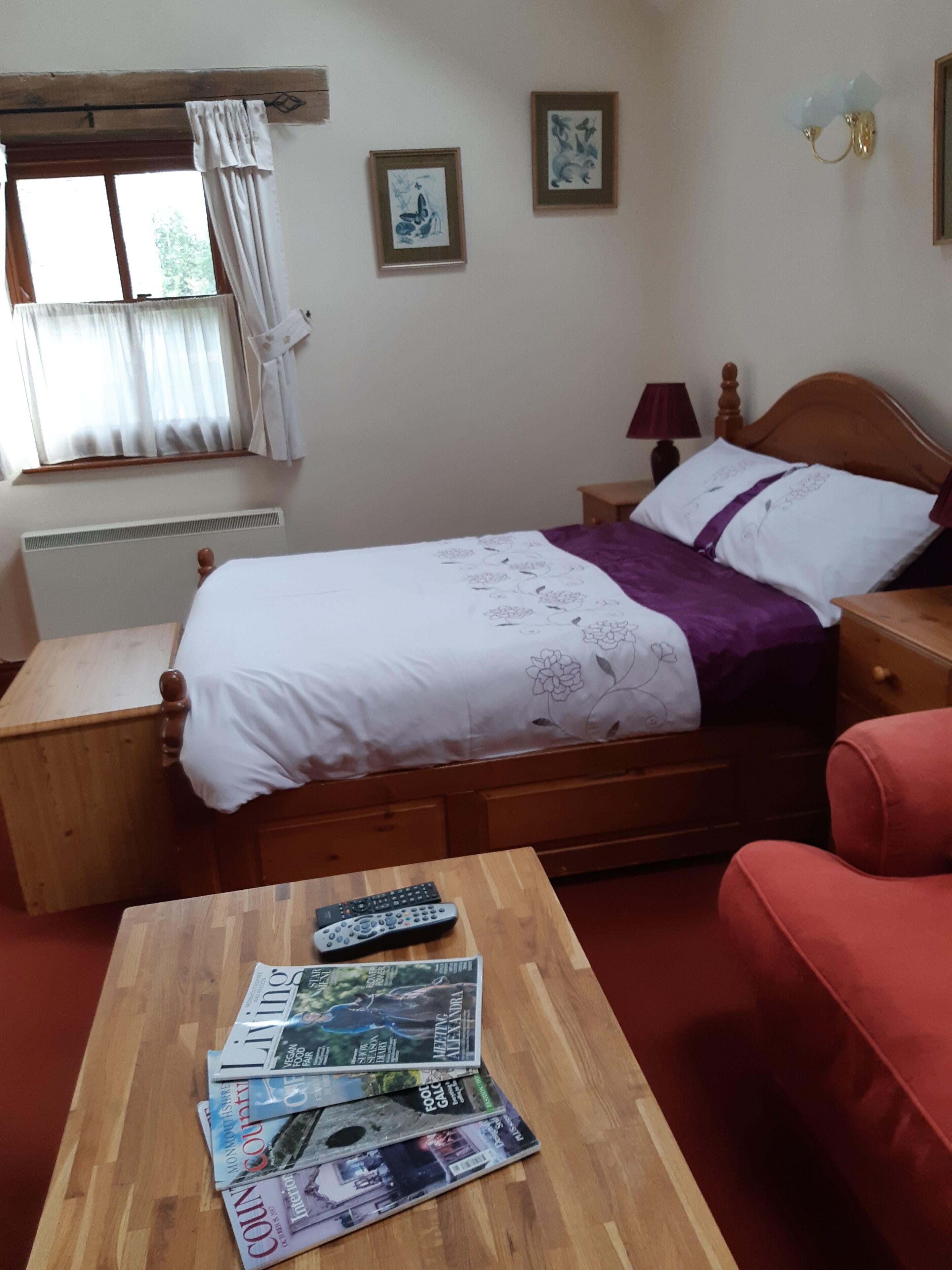 A bedroom with a wooden bed, a wooden nightstand with magazines, a red chair, and a window with blinds.