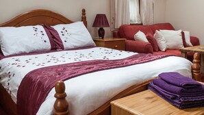 Un lit bien fait, recouvert d’une couvre-lit blanche et violette, une table de chevet en bois avec une lampe, un canapé rouge avec des coussins blancs et une table en bois sur laquelle sont pliées des serviettes violettes.