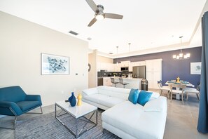Un salon moderne avec un canapé d’angle blanc, un fauteuil bleu et une table basse. On aperçoit une cuisine à l’arrière-plan, avec un coin repas.