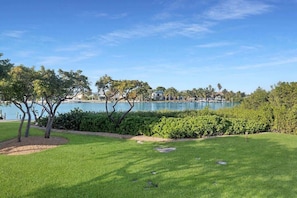 A well-maintained grassy area with a water body in the background, surrounded by trees and a hedge.
