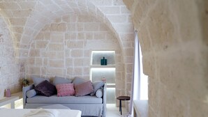A cozy room with a stone wall, a grey sofa with cushions, a small table, and a bed with a white blanket. - Vico Bianco (Ostuni)