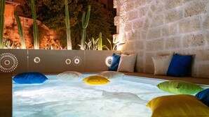 A room with a large, illuminated water feature in the center, a stone wall, and a seating area with cushions. - Vico Bianco (Ostuni)