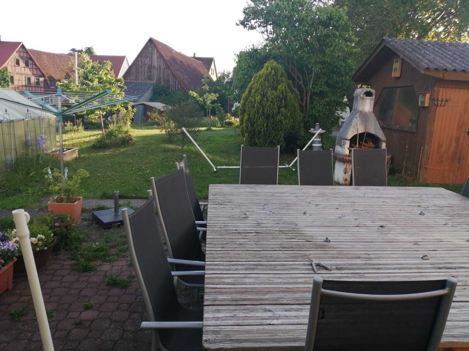 Un espace de restauration extérieur rustique, avec une table en bois et des chaises en métal, entouré d’un jardin où poussent différentes espèces de plantes et bordé d’un petit sentier en briques.