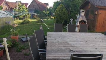Un espace de restauration extérieur rustique, avec une table en bois et des chaises en métal, entouré d’un jardin où poussent différentes espèces de plantes et bordé d’un petit sentier en briques.