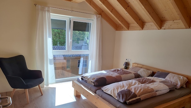 A bedroom with a wooden bed, a chair, and a balcony with a view of greenery.