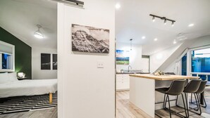 A modern, compact living space with a kitchenette, a dining area with bar stools, a bed with a striped rug, and a large window offering a view. - Coffee Lover`s Loft! Walk to Lake & brews. A/C (Estes Park)