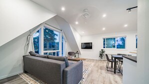 A modern loft-style room with a sloped ceiling, a gray sofa, a dining area, and large windows offering a view of trees. - Coffee Lover`s Loft! Walk to Lake & brews. A/C (Estes Park)