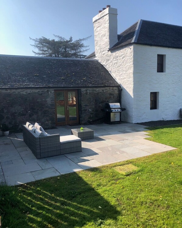A house with a stone exterior, a white chimney, a patio with a grill, and a grassy yard. - Comfortable Detached House with Mountain and Meadow views (Appin)