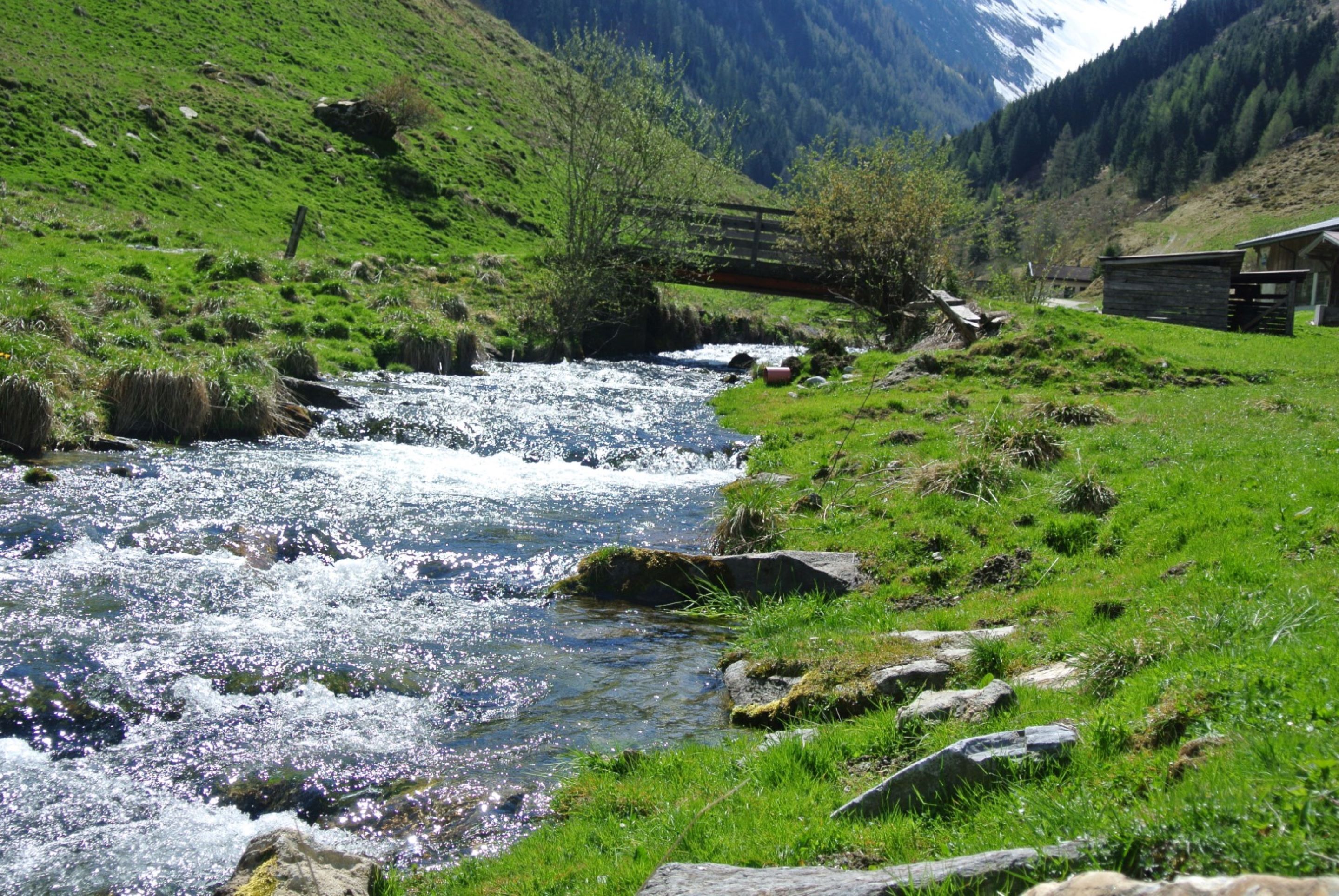 Un río que serpentea por un valle verde con un puente de madera y un edificio pequeño a la derecha.