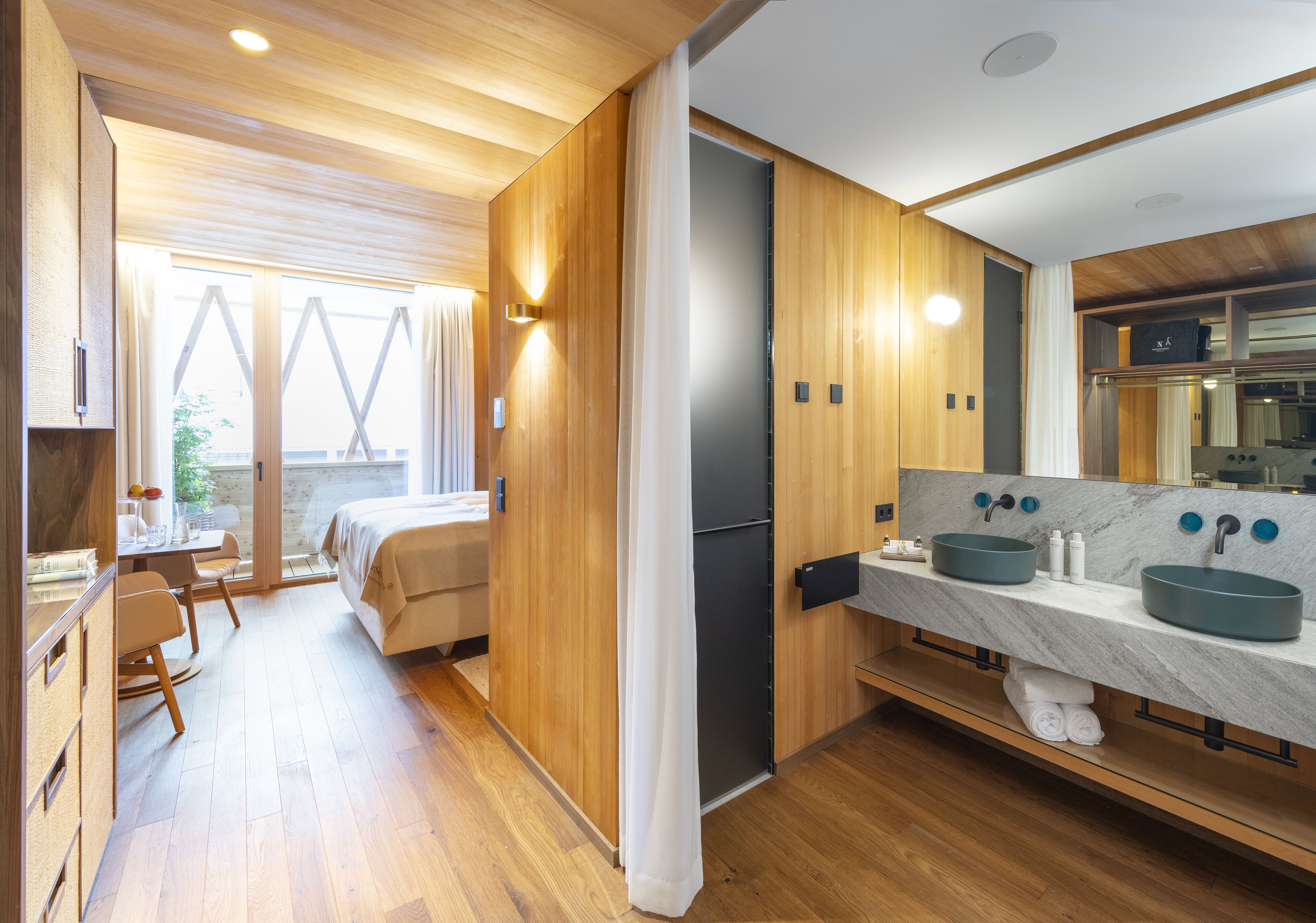 A modern hotel room with wooden flooring, a large bed, a desk with a chair, a bathroom with two sinks and a mirror, and a view through the window.