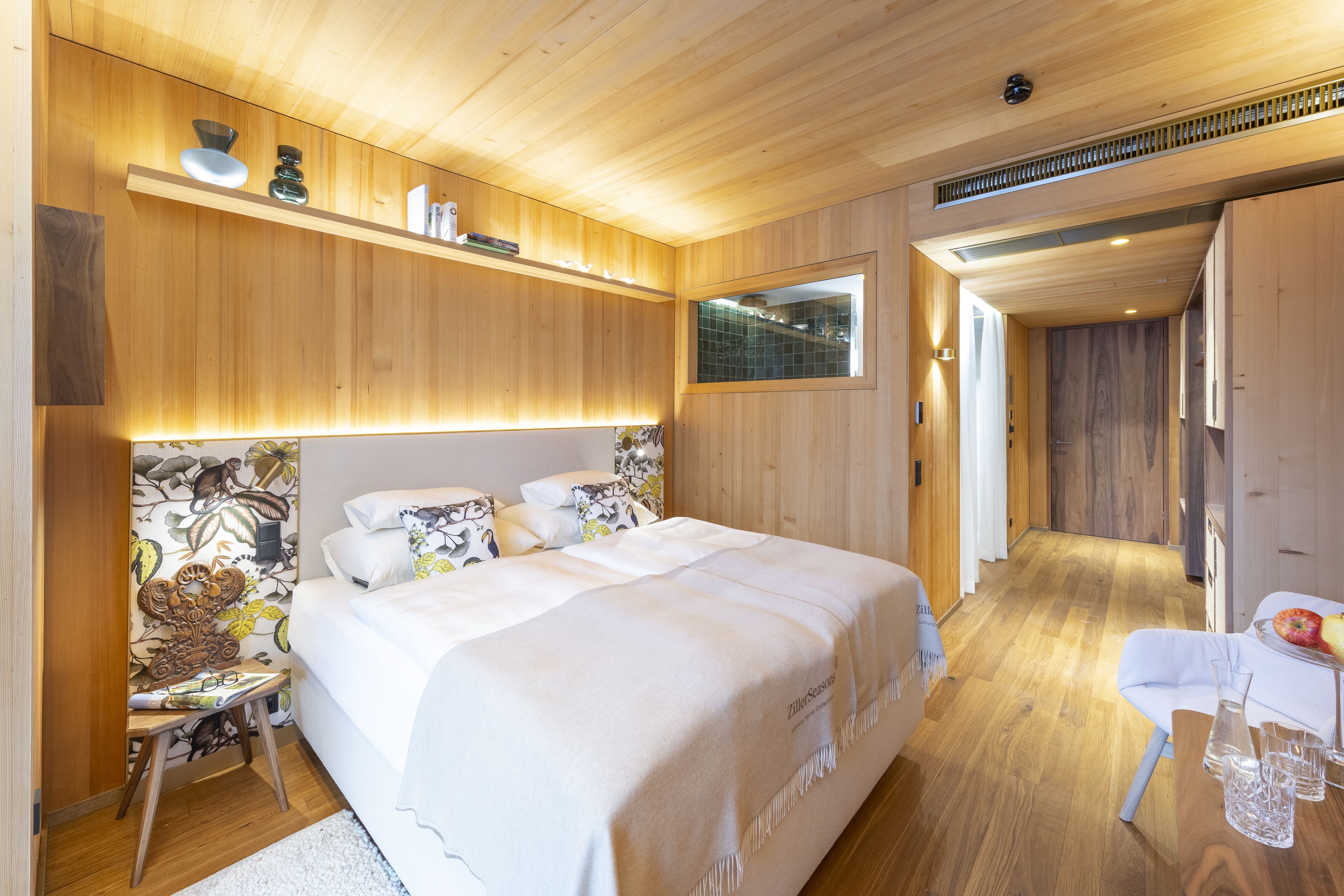 Un dormitorio moderno con techo de madera, una cama grande con ropa blanca, una mesita con un jarrón y vasos, y una silla.