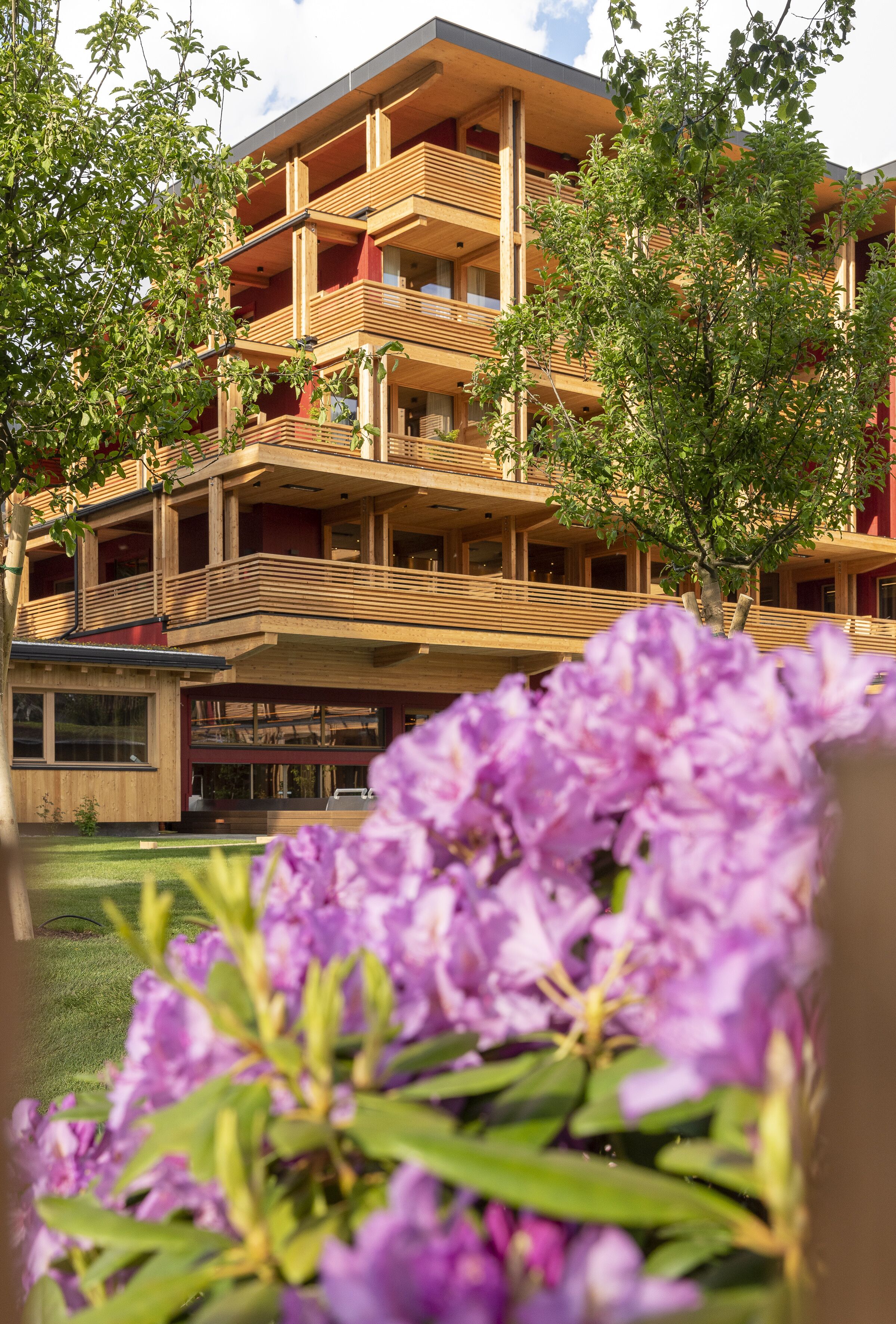A multi-story building with balconies and a garden area with purple flowers in the foreground.