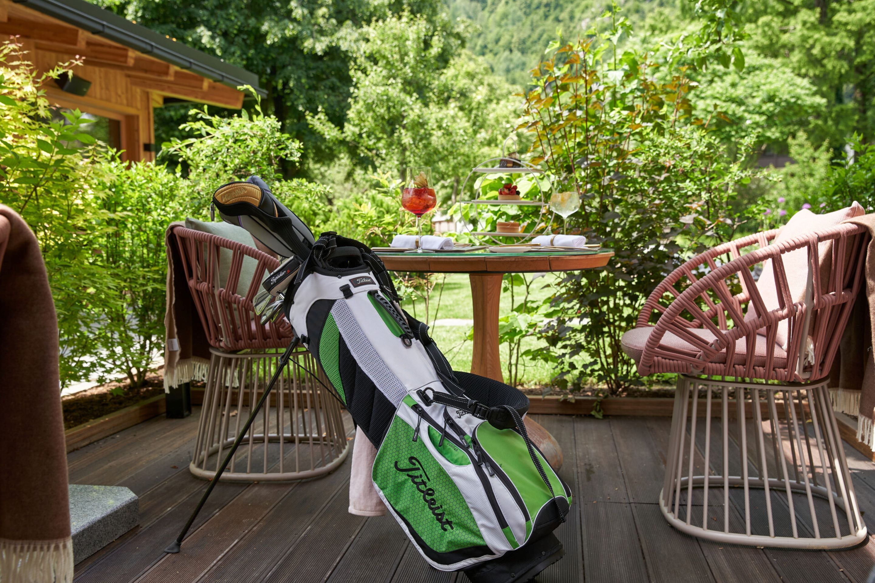 A golf bag on a wooden deck with wicker chairs, a table with a drink, and lush greenery in the background.