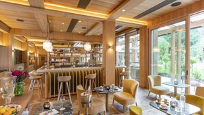 Eine moderne Bar mit Holzakzenten und einer Theke, auf der Flaschen und Gläser präsentiert werden. Gelbe Stühle und Tische sind zum Essen angeordnet, mit Blick auf Grünflächen im Außenbereich.