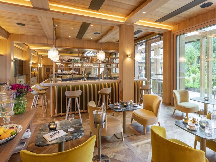 A modern bar area with wooden accents and a bar counter displaying bottles and glasses. There are yellow chairs and tables arranged for dining, with a view of greenery outside.