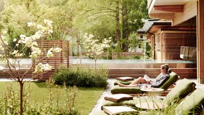 Eine Frau entspannt auf einem grünen Liegestuhl am Pool, mit einer Holzhütte und üppigem Grün im Hintergrund.