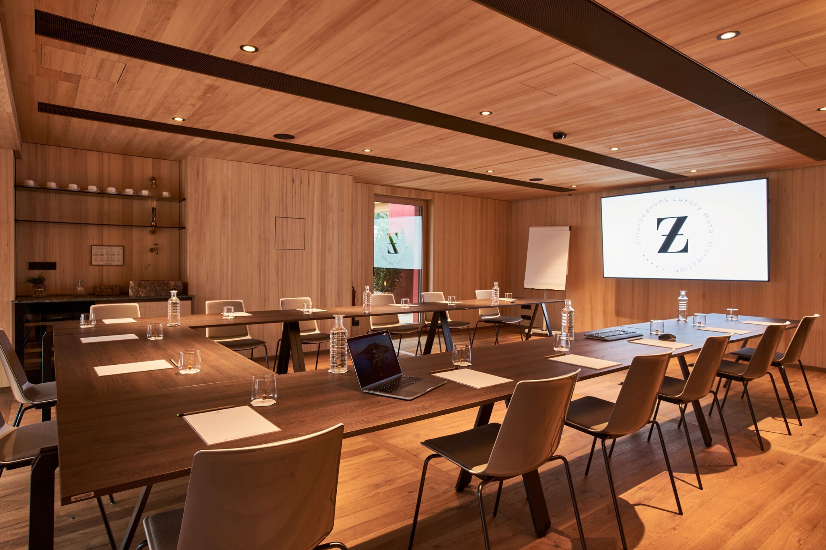 A conference room with a long wooden table, multiple chairs, and a projector screen displaying a logo.