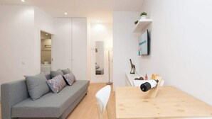 Un salon moderne avec un canapé gris, des chaises blanches, une table en bois et un téléviseur fixé au mur.