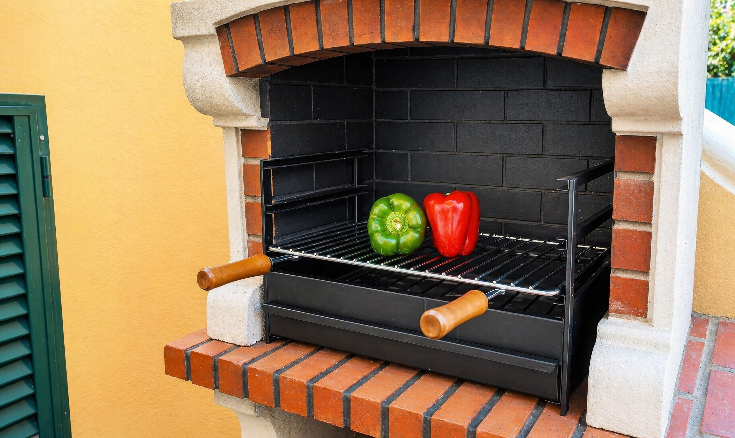 A brick-built outdoor grill with two wooden handles and two peppers on the grill grate.