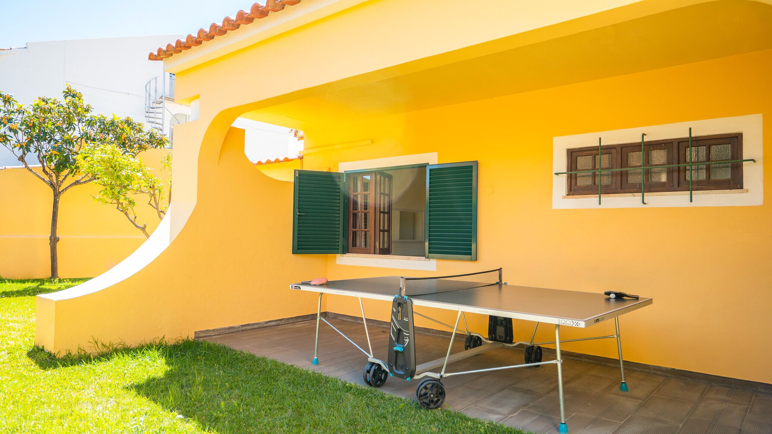 A yellow building with a ping pong table, a window with green shutters, and a small tree in the yard.