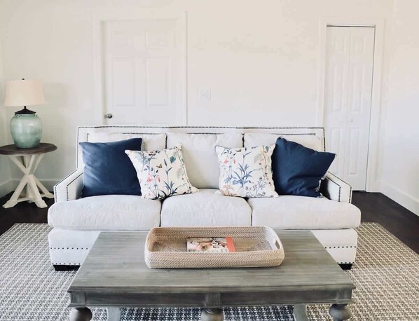 A white sofa with blue and floral cushions, a woven tray on a wooden coffee table, and a small round table with a lamp and vase.