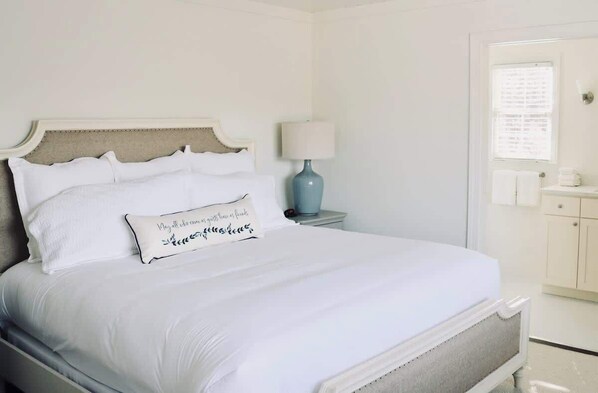 Una cama bien hecha con sábanas blancas y un cojín decorativo, una lámpara de noche y una ventana con persianas.