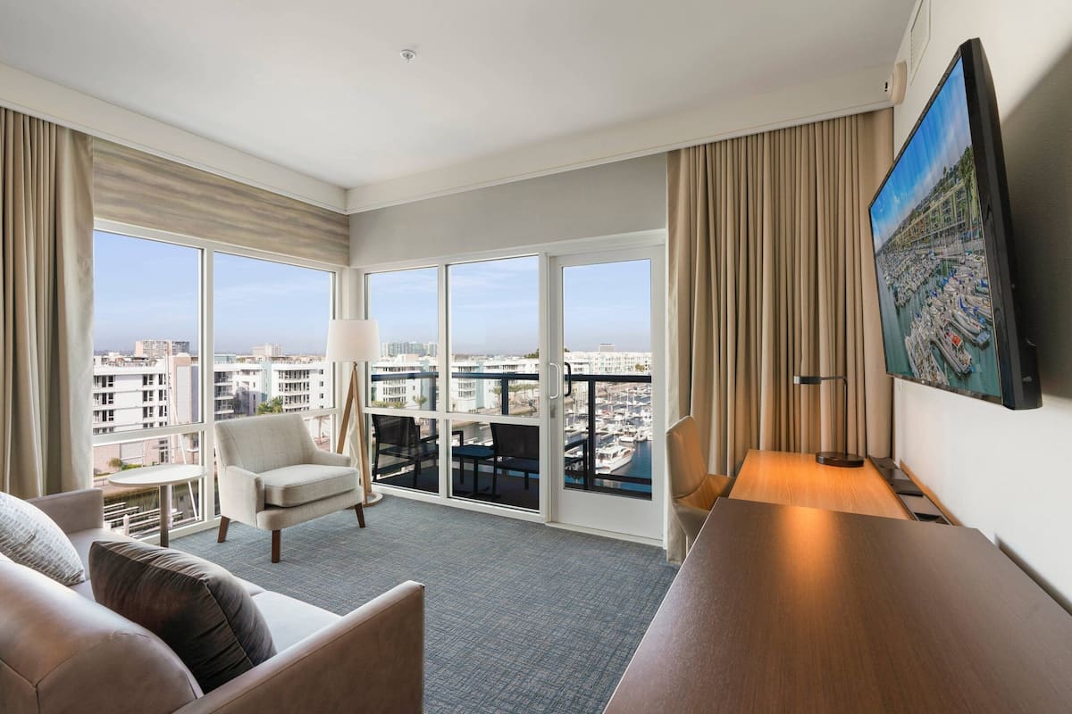 A hotel room with a large window, a flat-screen TV, a sofa, a chair, and a small table.