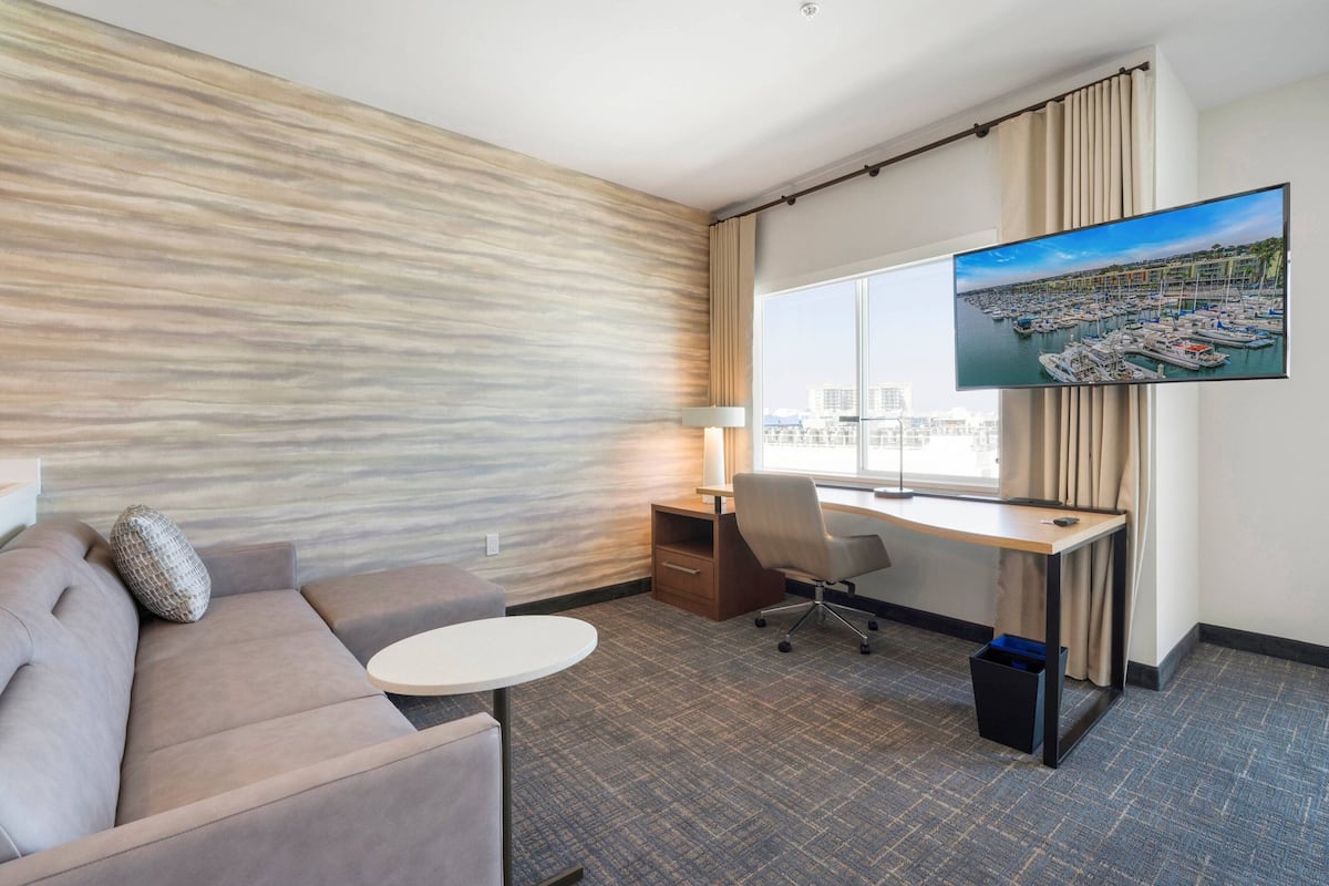 A hotel room with a large window, a flat-screen TV, a sofa, a desk, and a chair.
