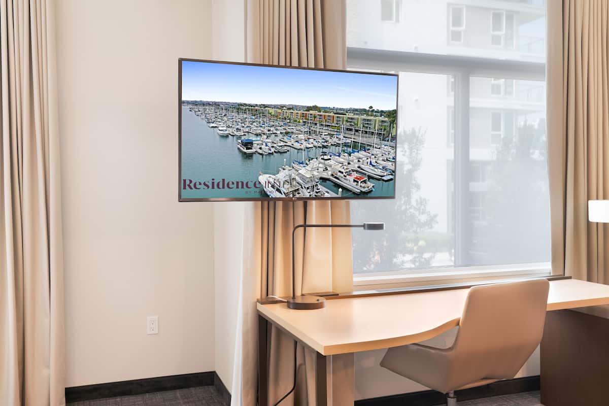 A hotel room with a desk, chair, and a TV mounted on the wall displaying a marina view.