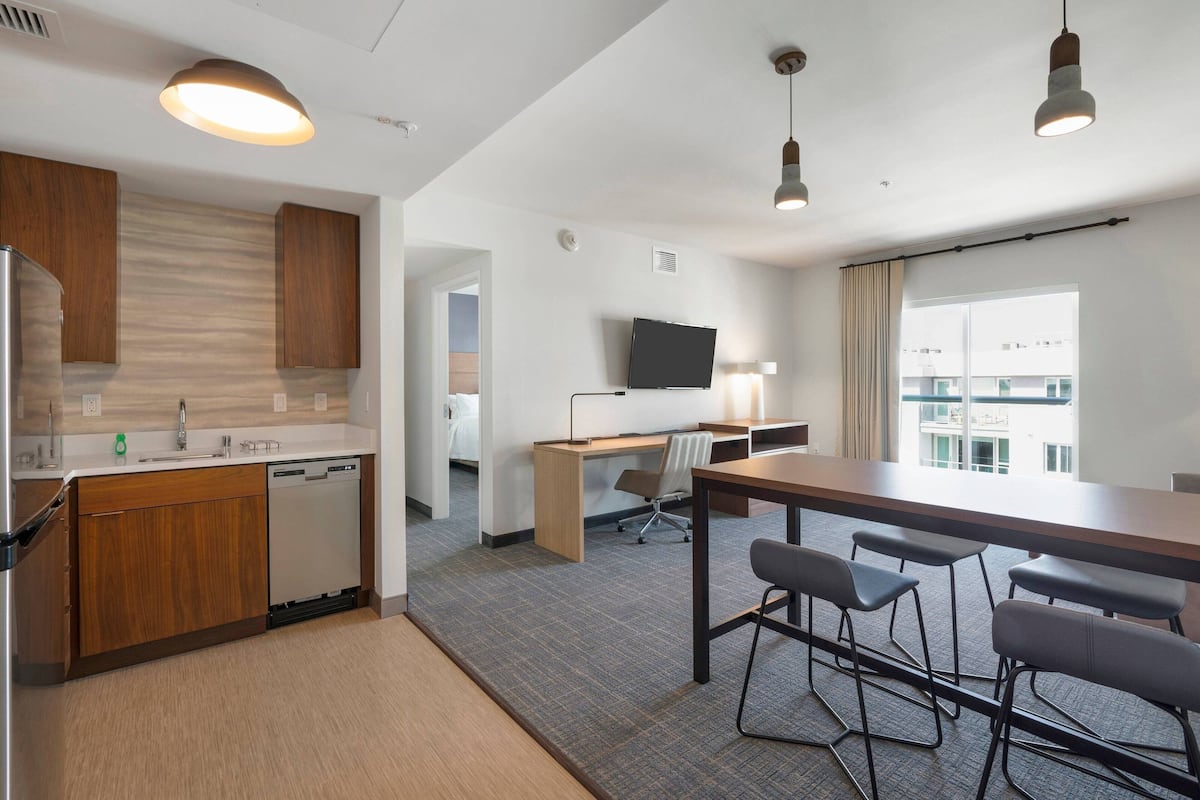 A modern hotel room with a kitchen, a dining area with a table and chairs, a desk with a chair, a television, and a large window with a view.