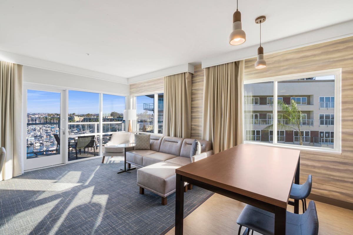 A modern hotel room with a large window offering a city view, a sofa, a dining table, and a balcony with a view of the marina.