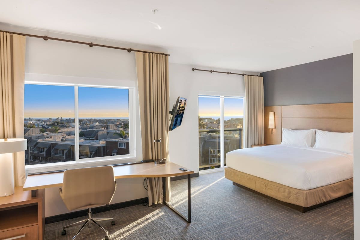 A hotel room with a large bed, a desk, a chair, and a view of the city through large windows.