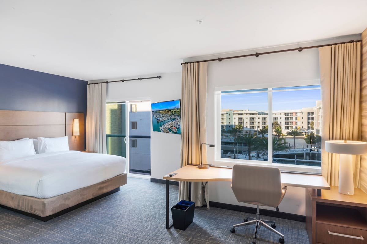 A hotel room with a large bed, a desk, a chair, a lamp, and a view of buildings through the window.