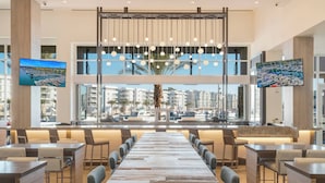 Um restaurante moderno com uma longa mesa de madeira, assentos no balcão e grandes janelas com vista para o porto e edifícios.