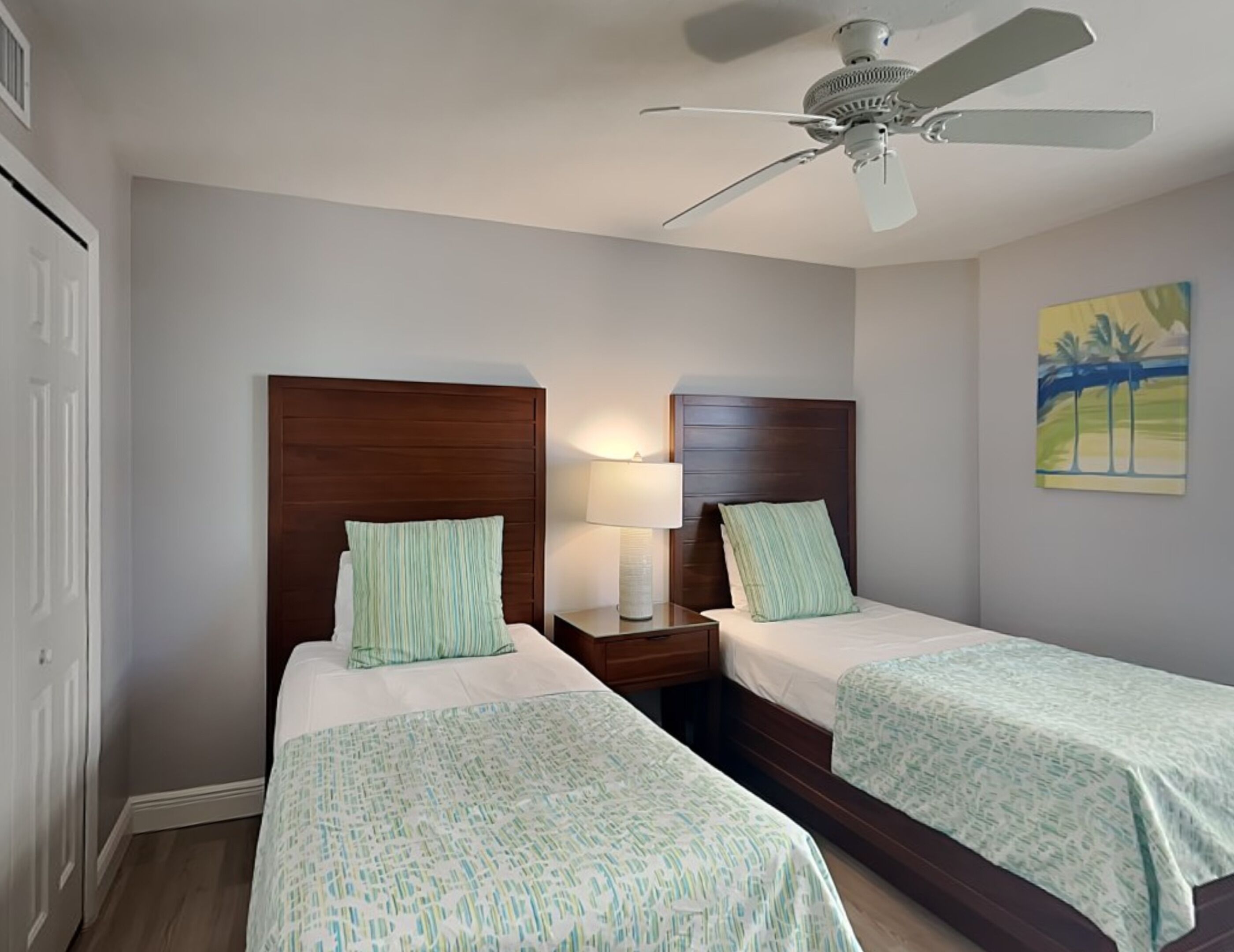 A hotel room with two single beds, a ceiling fan, a nightstand with a lamp, and a painting on the wall.
