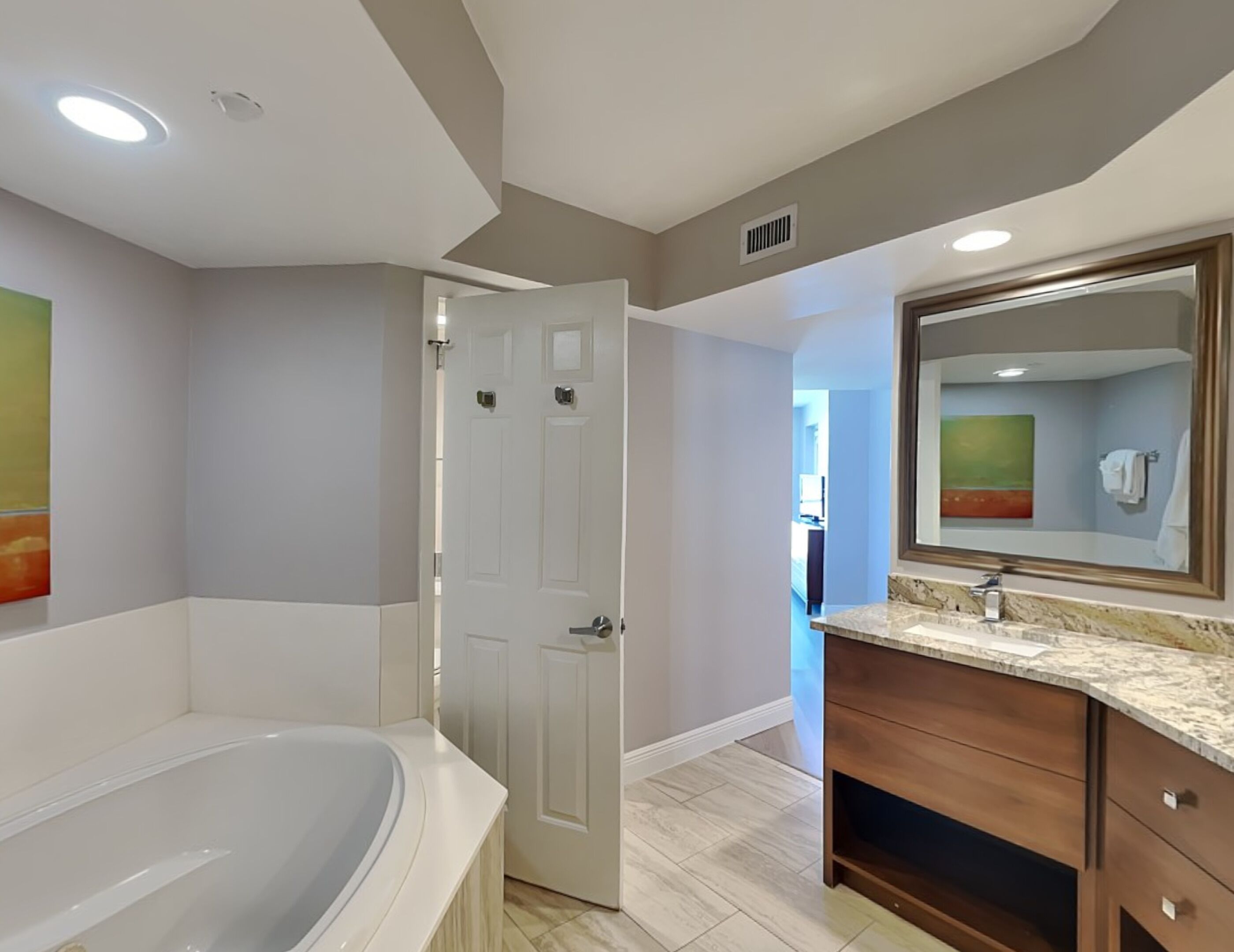 A bathroom with a large bathtub, a wooden vanity with a mirror, and a door leading to another room.