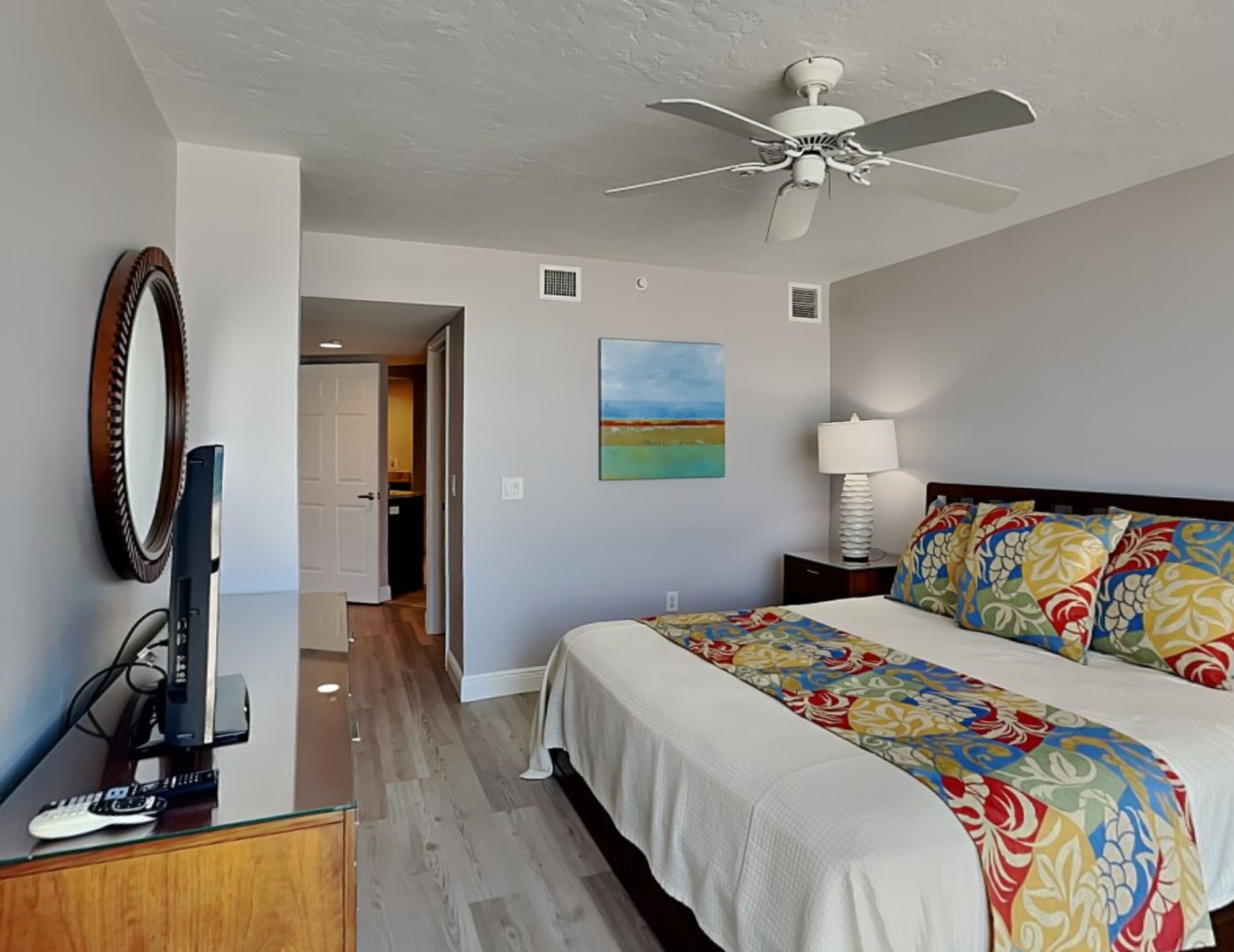 A hotel room with a bed, a ceiling fan, a TV, and a painting on the wall.