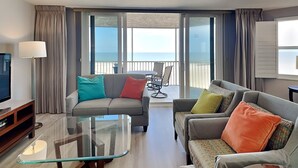 A modern living room with a sofa, armchairs, a glass coffee table, and a TV. There is a balcony with a table and chairs, and a view of the sea. - Gullwing 606 by Distinctive Beach Rentals (Fort Myers Beach)