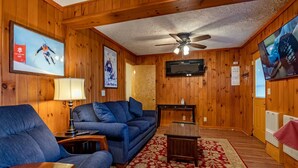 A cozy room with wooden walls, a blue sofa, a red floral carpet, a ceiling fan, and a TV on the wall.