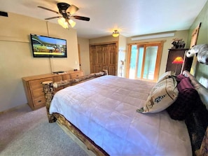 A bedroom with a bed, a wooden dresser, a ceiling fan, a television, and a door leading to another room. - Diamond Jewel - Pool table, Fire Pit (Deadwood)