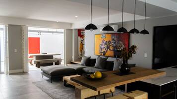 A modern living room with a sofa, a wooden dining table, hanging pendant lights, and a colorful abstract painting.