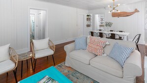 Una sala de estar con un sofá blanco, dos sillones de mimbre, una mesa de centro de vidrio y una zona de comedor con una mesa y sillas blancas.