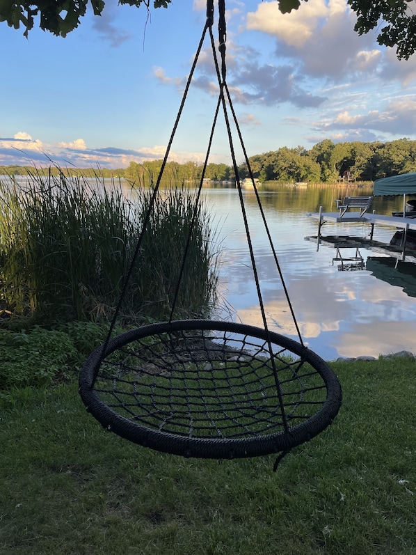 A hanging chair by a lake with a dock and boats in the background.