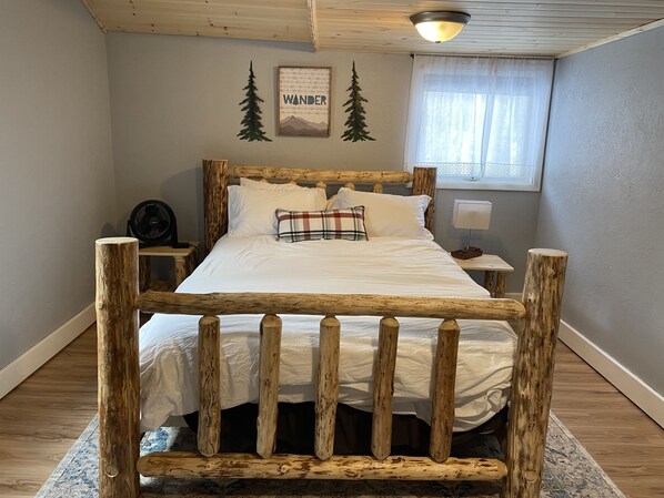A bedroom with a wooden bed frame, white bedding, a plaid pillow, a fan, a nightstand, a lamp, and a window with sheer curtains.