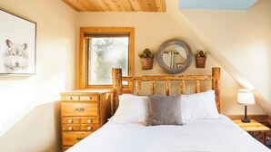 A bedroom with a wooden headboard, a dresser, a bedside table with a lamp, and a window with a view of trees.
