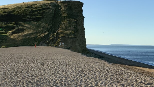 A beach with a steep cliff, a clear sky, and a calm sea. - Church Cottage (Bridport)