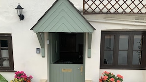 A green door with a small porch, two potted plants on either side, and a white wall with a window and a light fixture. - Church Cottage (Bridport)