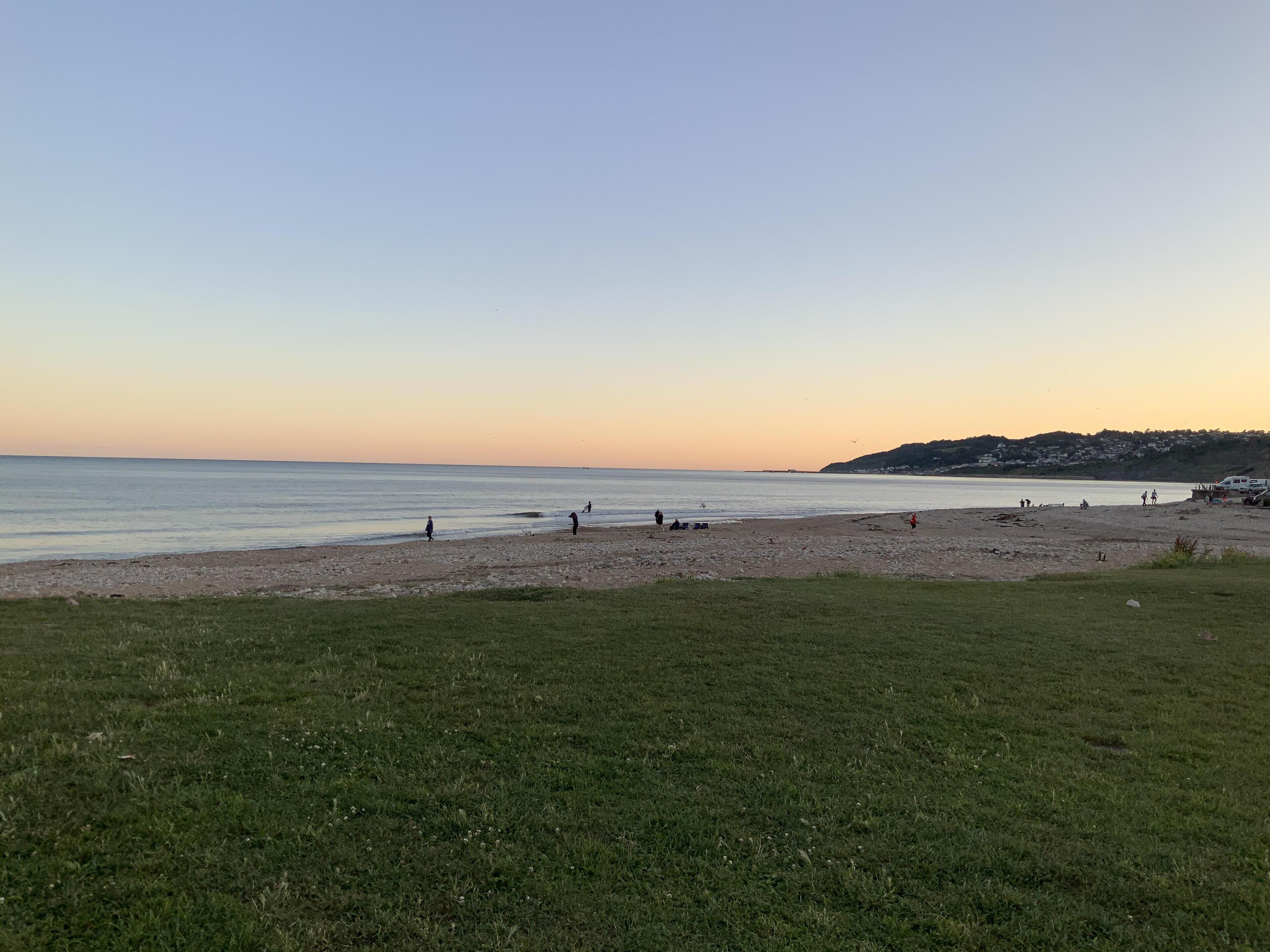 Une plage où l’on voit des gens se promener et jouer, une zone herbeuse au premier plan et un paysage vallonné en arrière-plan.