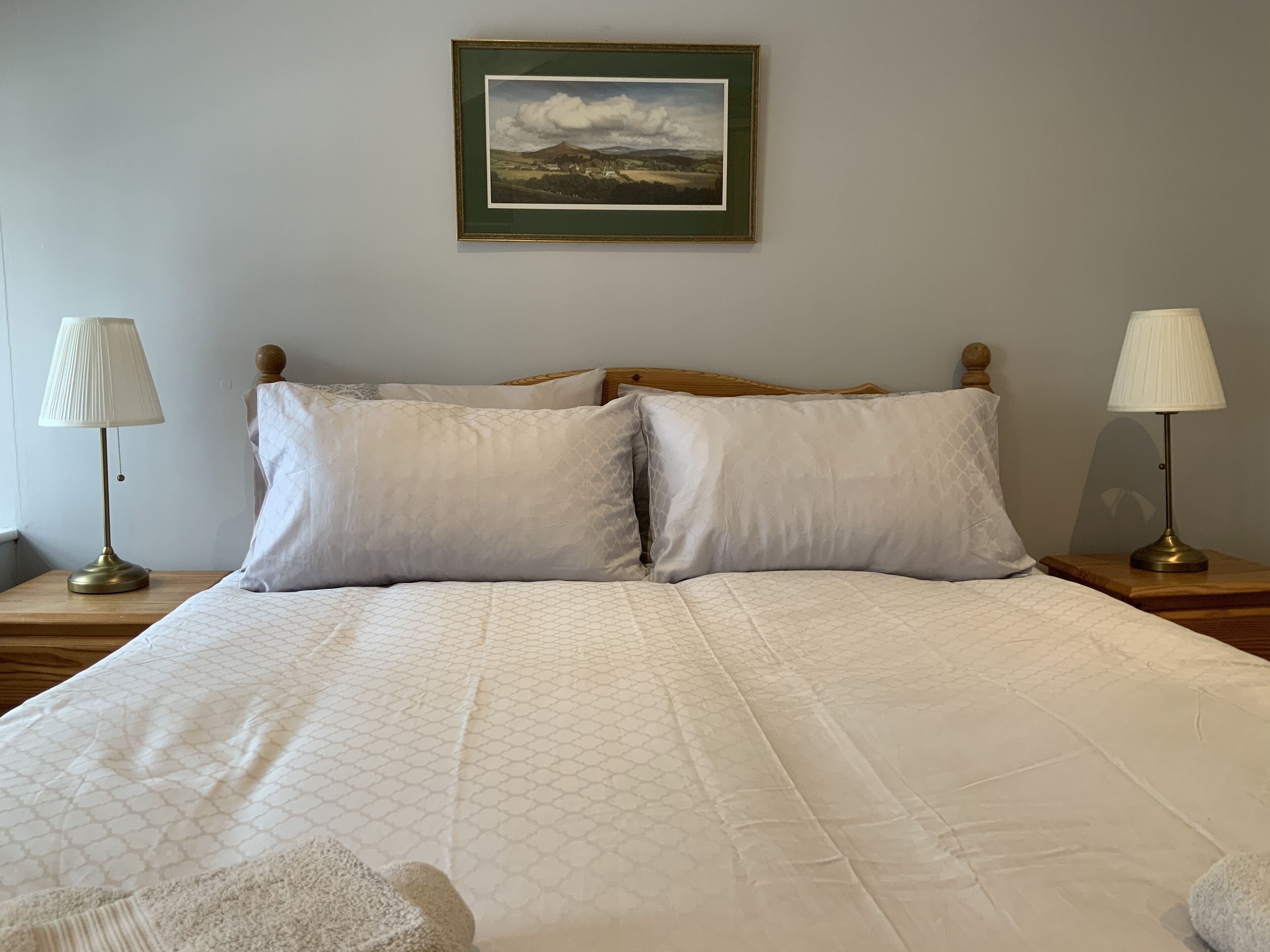 A neatly made bed with a quilted pattern, two bedside lamps, and a framed landscape painting on the wall.