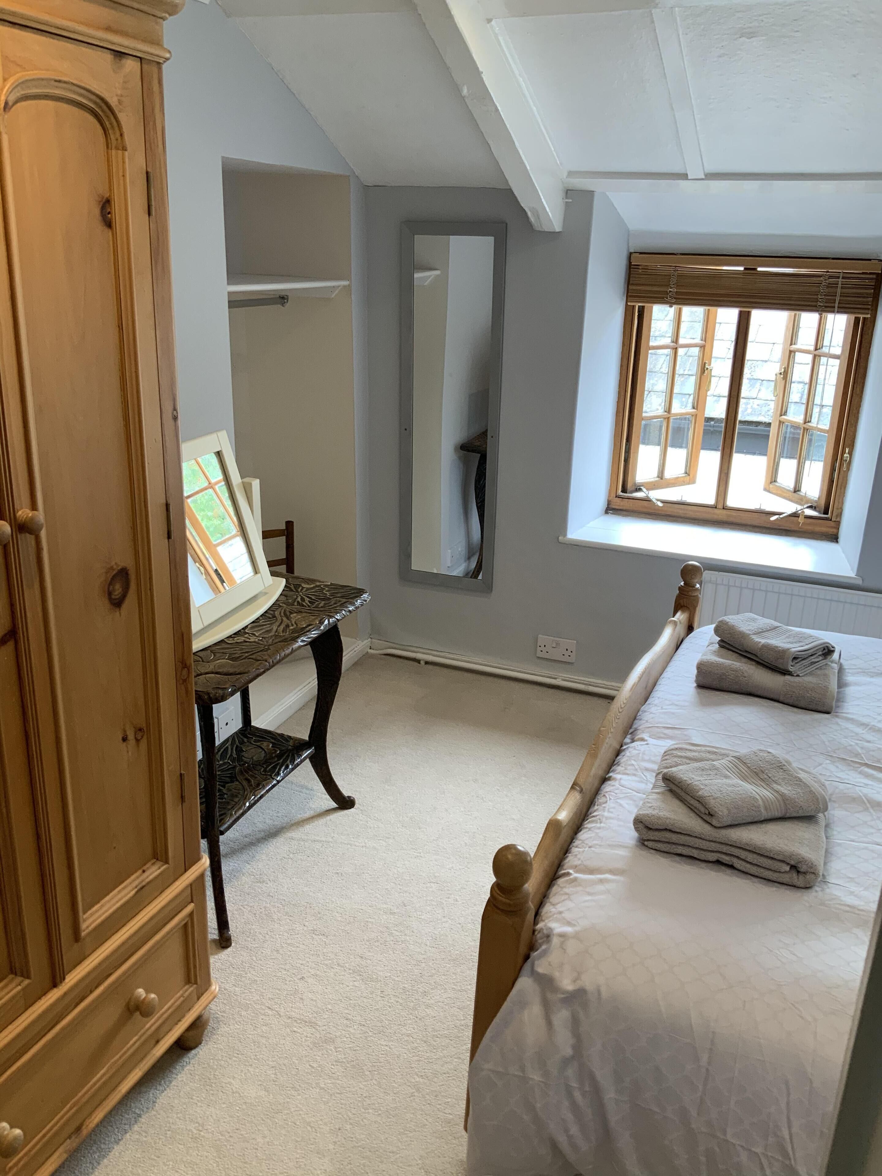 A bedroom with a bed, a wooden wardrobe, a desk with a mirror, and a window with blinds.